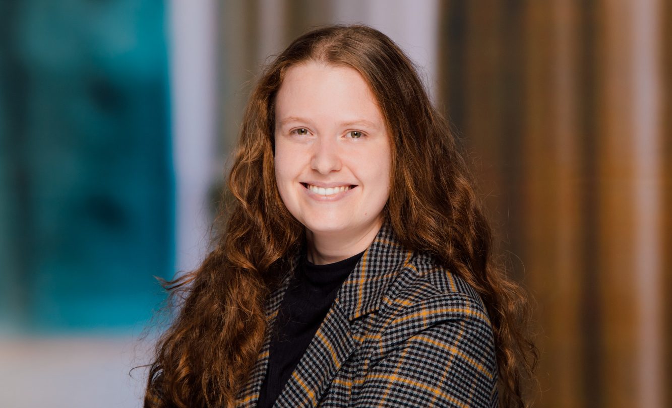 Judith Thomann Junge Frau mit langen, lockigen Haaren, lächelnd in einem modernen, hellen Raum.