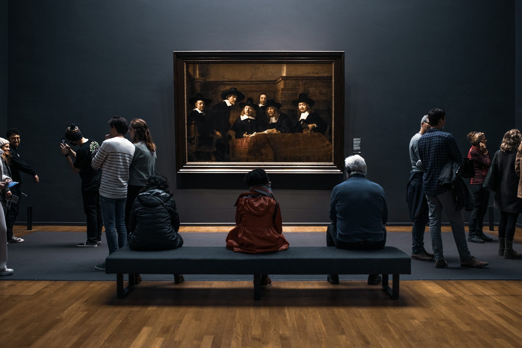 Besucher in einem Kunstmuseum betrachten ein Gemälde vor einer dunklen Wand.